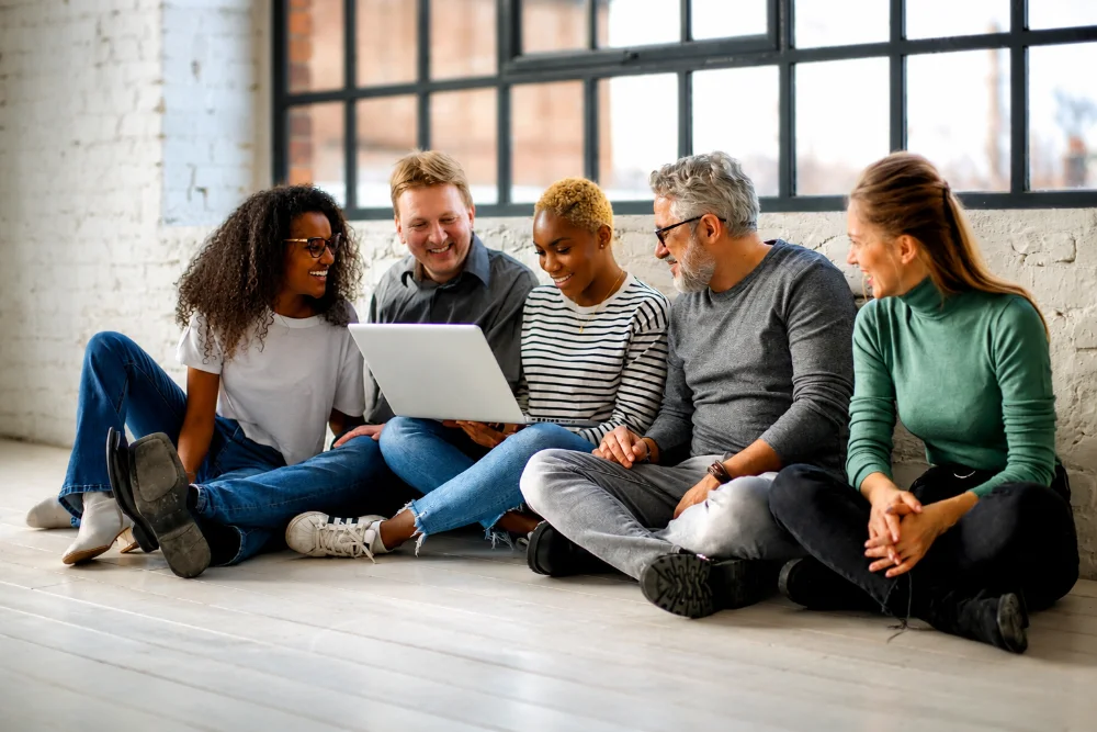 Five coworkers of different ages sitting together on the floor, smiling as they look at one laptop